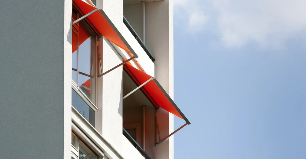 Markisen sind ausgefahren und hängen über den Fenstern eines mehrstöckigen Gebäudes an einer hellen Außenwand. Der Himmel ist blau mit wenigen Wolken.