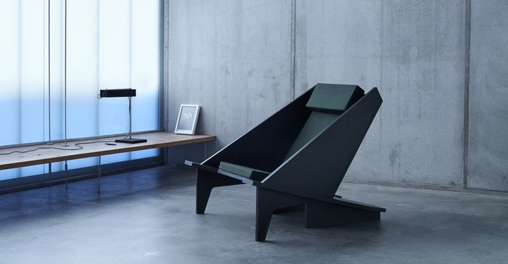 Ein moderner, kantiger Stuhl steht in einem minimalistischen, grauen Raum mit Betonwänden; links eine Holzbord mit Tischlampe und Bildrahmen vor einer matten Glaswand.