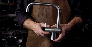 Eine Person mit schwarzem Hemd und brauner Lederschürze hält einen modernen silbernen Wasserhahn in beiden Händen in einer Werkstattumgebung mit Maschinen im Hintergrund.