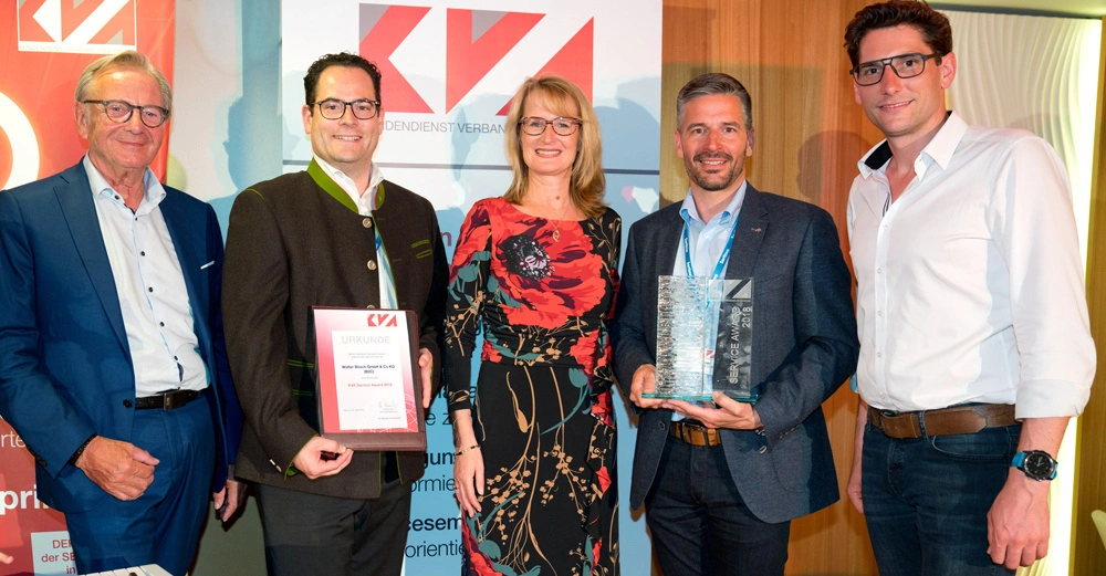 Fünf Personen lächeln und posieren für ein Foto in einem geschlossenen Raum einige halten eine Urkunde und eine Glasstatue KVA-Logo ist im Hintergrund zu sehen. Text: Urkunde Victor-Klein Sakko GmbH.