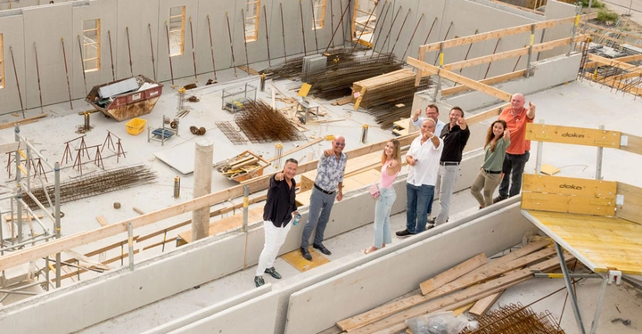 Galerie-Bild 3: Mehrere Menschen stehen auf einer Baustelle lächelnd und zeigen nach oben umgeben von Baugerüsten Baumaterialien und halbfertigen Betonstrukturen.