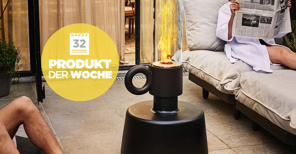 Ein schwarzer Ofen ist neben einer sitzenden Person, die in einem Zeitung liest, auf einer Terrasse platziert. Auf dem Bild steht in einem gelben Kreis die folgende Text: 32 PRODUKT DER WOCHE.