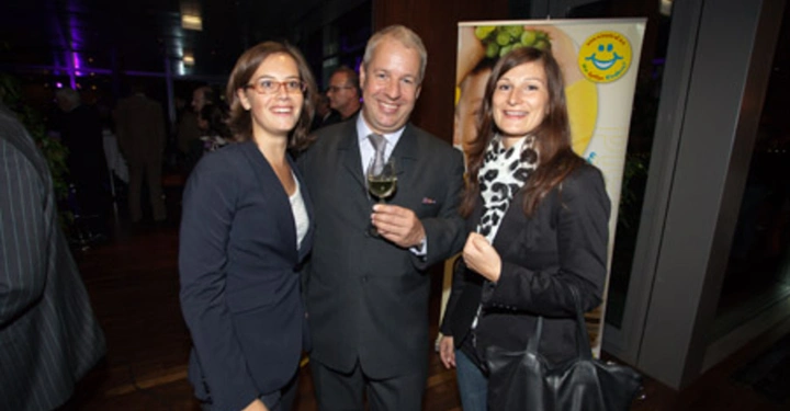 Galerie-Bild 76: Drei Personen stehen zusammen lächelnd in einem Raum. Eine hält ein Glas Wein. Hinter ihnen sind andere Menschen und ein Werbebanner mit einem lächelnden Gesicht und Trauben.