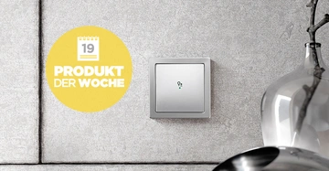 Ein silberner Lichtschalter ist an einer grauen Betonwand angebracht. Links befindet sich ein gelber Kreis mit Text 19 PRODUKT DER WOCHE. Rechts steht ein Silberkrug auf einem Tisch.