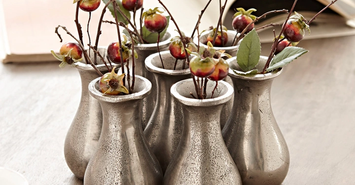 Galerie-Bild 7: Mehrere silberne Vasen halten Zweige mit kleinen roten Früchten und Blättern auf einem Tisch in einem Innenraum mit einem geöffneten Buch im Hintergrund.
