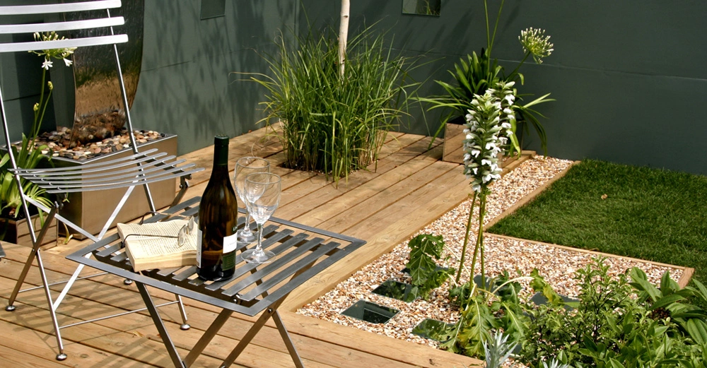 kleiner Metallklapptisch mit Metallklappstuhl auf Bodenplatten aus Holz im Hinterhof. Heller Kies, kurzer Rasen, alles eng aber schön