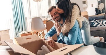 Mann schaut in Umzugskarton und lächelt während eine Frau ihn von hinten umarmt und küsst Wohnzimmer mit Kartons und Möbeln im Hintergrund