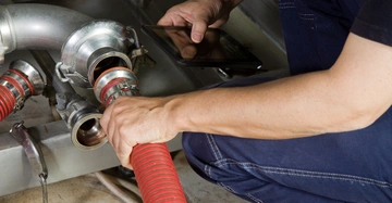 Ein Mensch in Arbeitskleidung hält eine rote Schlauchverbindung und benutzt ein Tablet in einer technischen Umgebung mit Metallrohren und Armaturen.