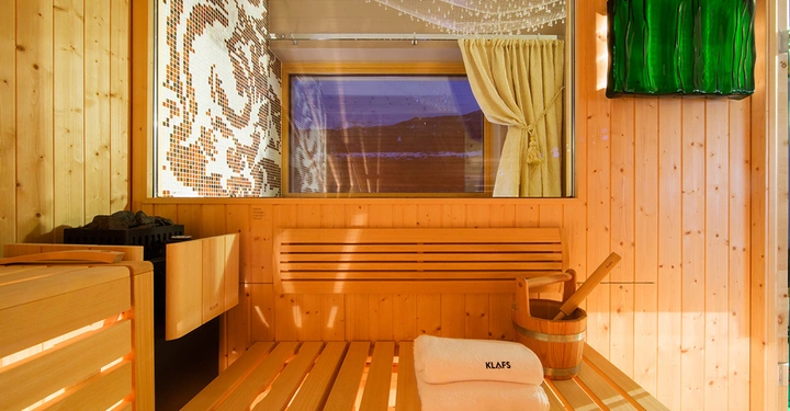 Saunaholzbank haltet zwei gefaltete Handtücher mit der Aufschrift KLAFS und einen Eimer mit Kelle daneben grosse Fenster bieten Blick nach draussen moderne Wandkunst gestaltet den Kontext