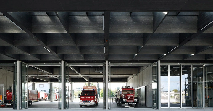 Galerie-Bild 11: Mehrere rote Feuerwehrautos parken in einer modernen Feuerwache mit schwarzen Deckenraster und Glaswänden in einer städtischen Umgebung. Auf den Fahrzeugen steht "FEUERWEHR".