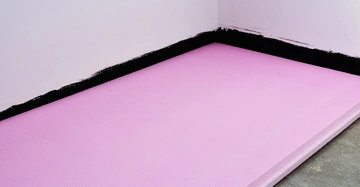 Rosa Dämmplatte liegt auf grauem Boden in einer Ecke umgeben von weißen Wänden mit schwarzem Streifen entlang der Kante