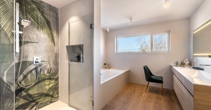 Galerie-Bild 2: Dusche wäscht Hände Badezimmer im modernen Stil mit Glasdusche Wand mit Blattmuster Holzfußboden weiße Badewanne mit Blick aufs Fenster grünem Stuhl Waschtisch mit beleuchtetem Spiegel Schriftzug LAMURISTA
