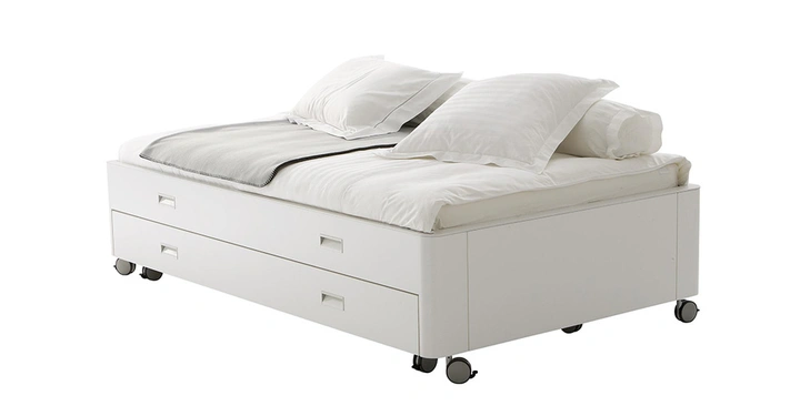Weißes Bett mit vier Kissen und Decke auf zwei großen Schubladen mit Griffen stehend auf kleinen schwarzen Rollen in einem neutralen weißen Raum