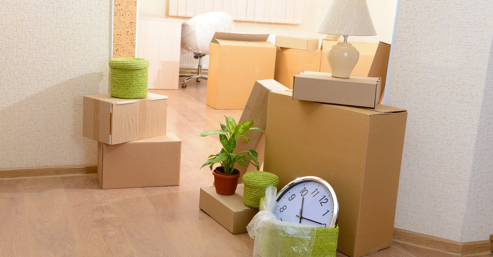 Pappkartons und Haushaltsgegenstände wie Pflanzen, eine Lampe und eine Uhr stehen in einem leeren Raum auf einem Holzboden und vor einer beige Tapete und Fenstern mit Jalousien.