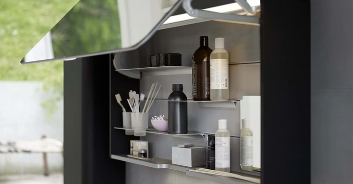 Im geöffneten Spiegelschrank stehen verschiedene Hygieneartikel auf Regalböden. Im Hintergrund ist ein grüner Außenbereich zu sehen.