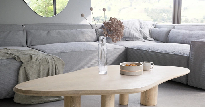 Ein hölzerner Kaffeetisch mit einer Vase und Schale darauf steht vor einem grauen Sofa in einem modern eingerichteten Wohnzimmer mit großen Fenstern und Blick auf hügelige Landschaft
