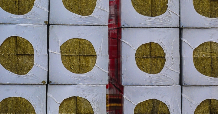 Anordnung von beigen Steinwollblöcken verpackt in weißer Plastikfolie mit runden Ausschnitten, gestapelt nebeneinander in einer symmetrischen Struktur, rotes Klebeband in der Mitte.