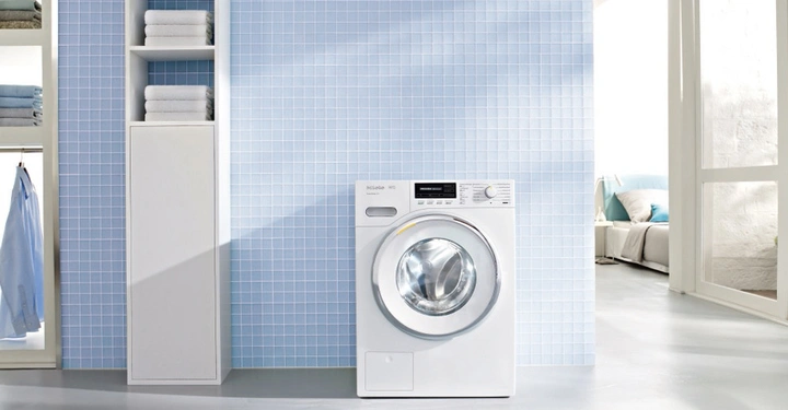 Waschmaschine stehend vor einer hellblauen gefliesten Wand daneben Regalfächer mit gefalteten Handtüchern im modernen Waschraum viel natürliches Licht links Kleidung rechts Blick ins Schlafzimmer