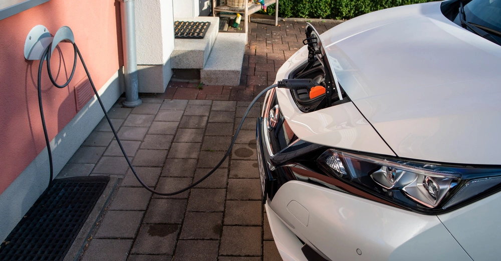 Ein weißes Elektroauto wird an einer Ladestation auf einem gepflasterten Weg vor einem Haus aufgeladen Kabel ist mit der Ladestation an der Wand verbunden und führt zur Autohaube.