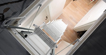 Leiter entfaltet sich in den Raum darunter mit Holzboden und einer geschlossenen Tür, Wände in hellem Ton, erkennbar ist ein Teil eines Stuhls und einer Lampe.