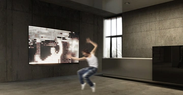 Person tanzt in einem modernen Raum mit grauen Betonwänden. Große beleuchtete Wandanzeige und Fenster im Hintergrund. Raum hat eine minimalistische Einrichtung.