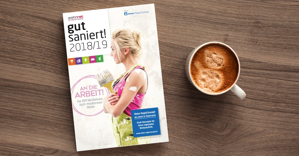 Eine Zeitschrift mit dem Titel "gut saniert! 2018/19" liegt auf einem Holztisch neben einer Tasse Cappuccino. Das Cover zeigt eine Frau mit einem Pinsel in der Hand. Text: "AN DIE ARBEIT! Im DIY-Verfahren zum modernen Heim"