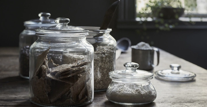 Galerie-Bild 1: Glasbehälter mit Deckeln stehen auf einem Holztisch und enthalten Lebensmittel wie Kekse Haferflocken und Mehl Im Hintergrund sind ein Fenster mit Pflanzen und eine Schaufel zu sehen