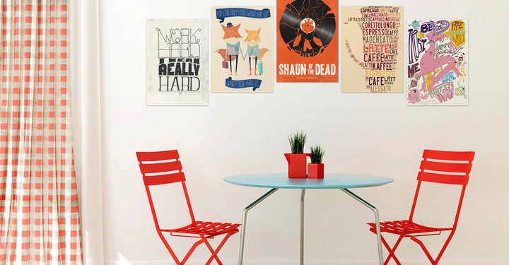 Runder Tisch mit zwei roten Stühlen und kleinen Pflanzen drauf vor einer weißen Wand mit fünf Postern mit verschiedenen Designs und Texten Work Hard then Really Hard You can only love what you can Shaun of the Dead sowie Kaffeetypen und einem bunten Design