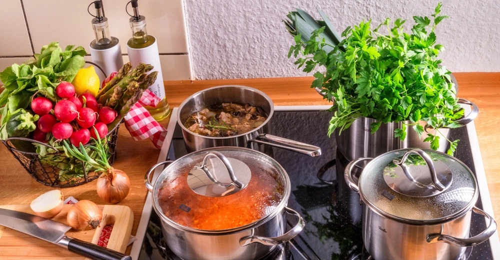 Kochtöpfe stehen auf einem Herd und köcheln Speisen In einem Korb daneben liegen Gemüse und Gewürze Küchenutensilien wie Messer und Schneidebrett sind ebenfalls auf der Arbeitsplatte zu sehen.