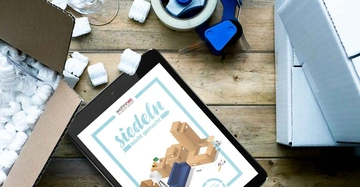 Tablet mit einem Umzugsartikel auf dem Bildschirm liegt neben einer geöffneten Kiste mit Verpackungsmaterial einem Klebebandabroller und einer weinglasähnlichen Schale auf einem Holzboden. Text auf dem Bildschirm wohnnet siedeln leicht gemacht Die besten Tipps für einen problemlosen Umzug.