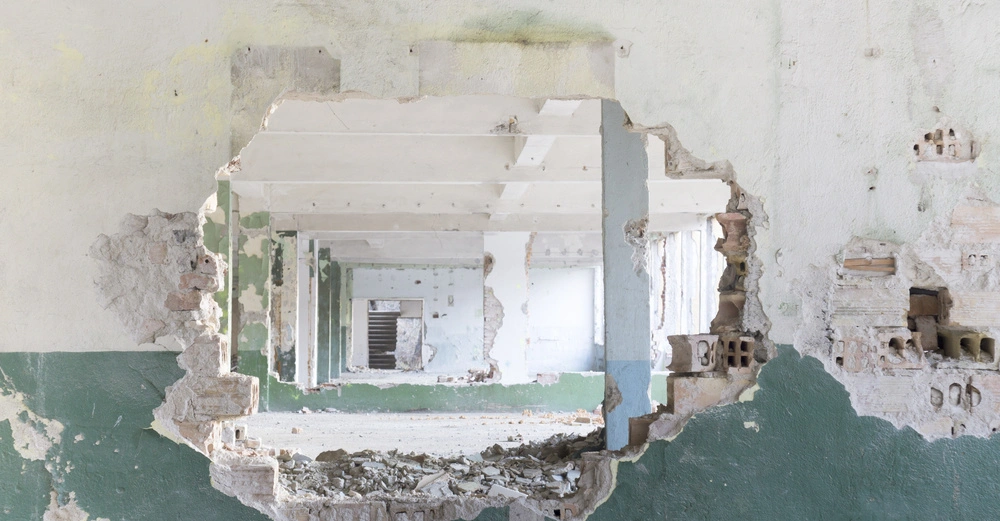 Ausgebrochene Mauer mit vielen Löchern führt zu leeren, heruntergekommenen Räumen hintereinander, zerbrochene Ziegelsteine und Schutt auf dem Boden, grün-weiße Wandfarbe teilweise abgeblättert in einem verlassenen Gebäude.
