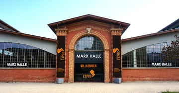Backsteingebäude mit großer Glasfront markiert als Marx Halle ein Willkommensschild und Expo-Plakate hängen; umliegender Bereich gepflastert und strukturiert. Text: MARX HALLE WILLKOMMEN frauenkraft EXPO 2020 EXPO DIE INNOVATIVE FACHMESSE FÜR SOZIALE BERUFE UND SOZIALE DIENSTLEISTUNGEN