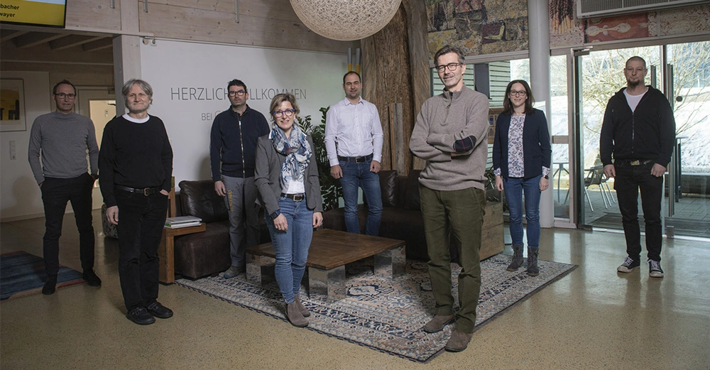 Menschen stehen in einem Raum vor einer Wand mit der Aufschrift HERZLICH WILLKOMMEN BEI, einige sitzen auf braunen Sofas um einen niedrigen Holztisch, Hintergrund mit Pflanzen und Fenstern.