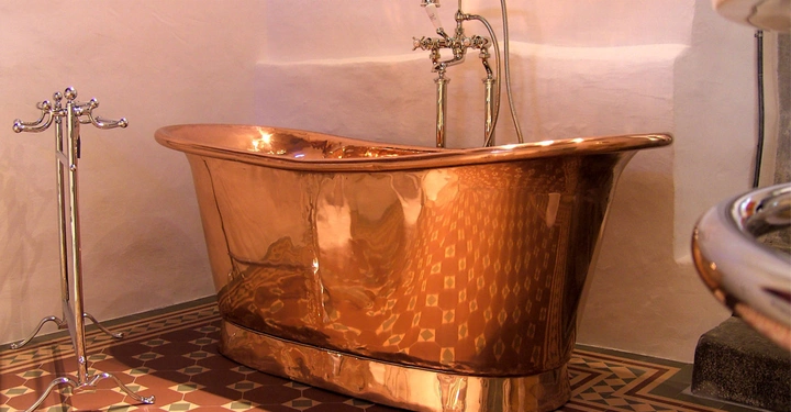 Kupferbadewanne steht still auf einem gemusterten Fliesenboden in einem Badezimmer mit beigen Wänden. Daneben ein verchromter Handtuchhalter und eine Wandarmatur mit zwei Wasserhähnen.