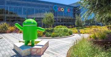 Eine große grüne Android-Statue steht auf bunten Würfeln vor dem Google-Hauptquartier mit verglaster Fassade und buntem Google-Logo im grünen Campusbereich.