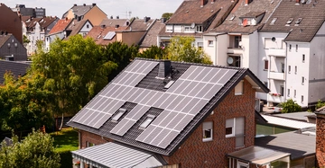 Ein Haus mit Solarpaneelen bedeckt das Dach steht in einem Wohngebiet mit mehrstöckigen Nachbargebäuden und umgeben von Gräsern und Bäumen.