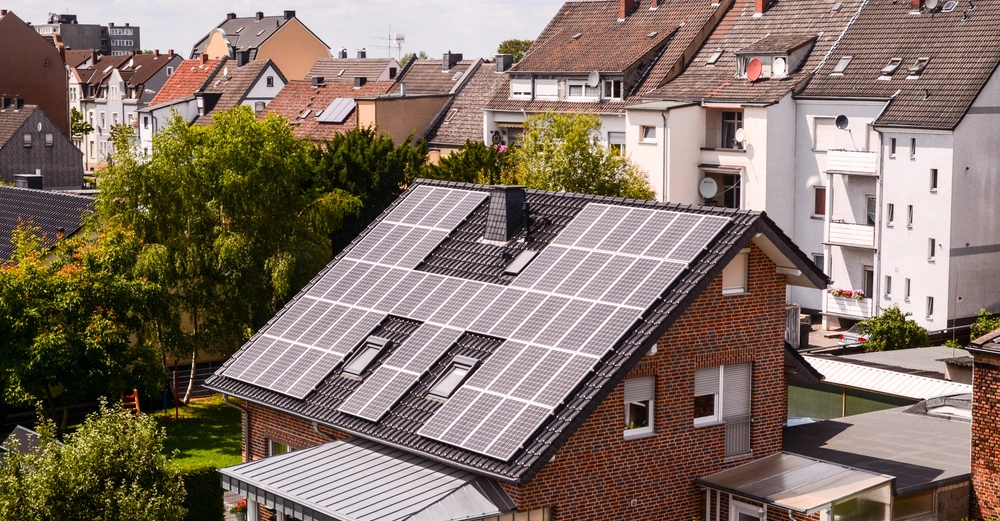 Ein Haus mit Solarpaneelen bedeckt das Dach steht in einem Wohngebiet mit mehrstöckigen Nachbargebäuden und umgeben von Gräsern und Bäumen.