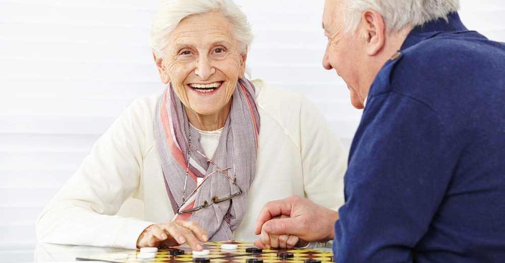 Ältere Frau mit Brille spielt Dame an einem Glastisch mit einem älteren Mann in einem hellen Raum. Sie lächelt, beide konzentrieren sich auf das Spiel.