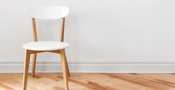 Weißer Holzstuhl steht auf hellbraunem Parkettboden vor einer weißen Wand in einem minimalistisch eingerichteten Raum.