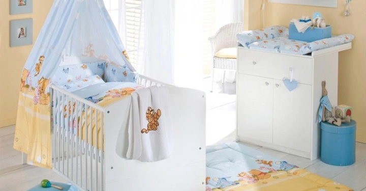 Galerie-Bild 7: Weißes Babybett mit farbigem Bettzeug und Netz baldachin, neben Wickeltisch mit Wickelauflage und dekorativen Elementen in einem hellen Raum mit gelben Wänden und weißen Möbeln.