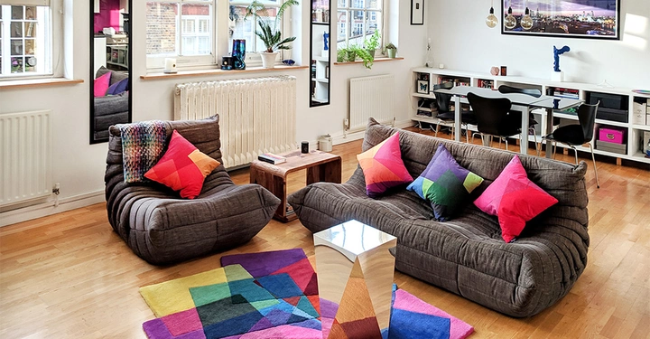 Grau gepolsterte Sofa und Sessel mit bunten Kissen auf einem Parkettboden vor einem Tisch in einem hellen Wohnzimmer mit Pflanzen Fenster und diversen Möbeln.