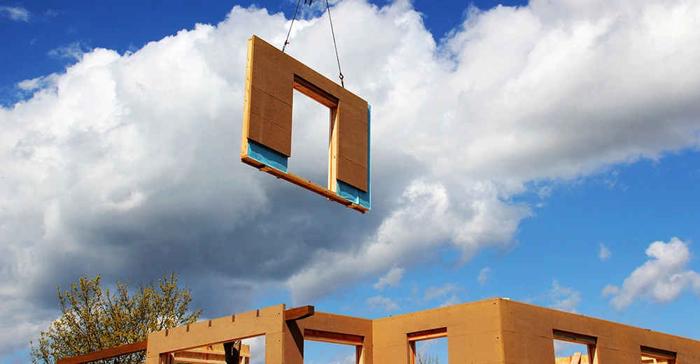 Blauer Himmel, Wolken, durch die Luft scheint ein Hausteil mit Türöffnugn aus Holz zu schweben. Fertighausbau at its best