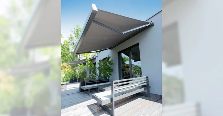 Markise erstreckt sich über Terrasse zwei graue Bänke stehen nebeneinander im Freien grüne Pflanzen umgeben den Bereich Hauswand im Hintergrund blauer Himmel