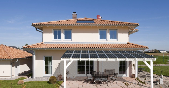 Ein zweistöckiges Haus mit roten Dachziegeln hat einen überdachten Außenbereich mit Solarpanelen im Garten. Im Vordergrund steht ein Gartenmöbelset auf einer gefliesten Terrasse.