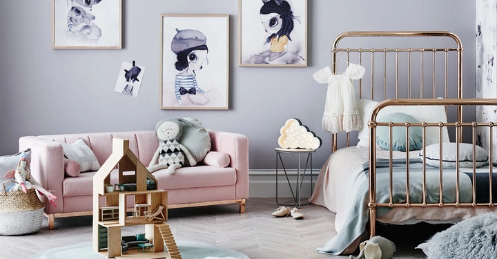 Holzpuppenhaus steht auf rundem Teppich in Kinderzimmer mit rosafarbenem Sofa und ausgestopftem Stofftier daneben im Hintergrund goldenes Gitterbett neben Tisch mit Wolkenlampe und weissem Kleid. Kunstbilder hängen an der Wand