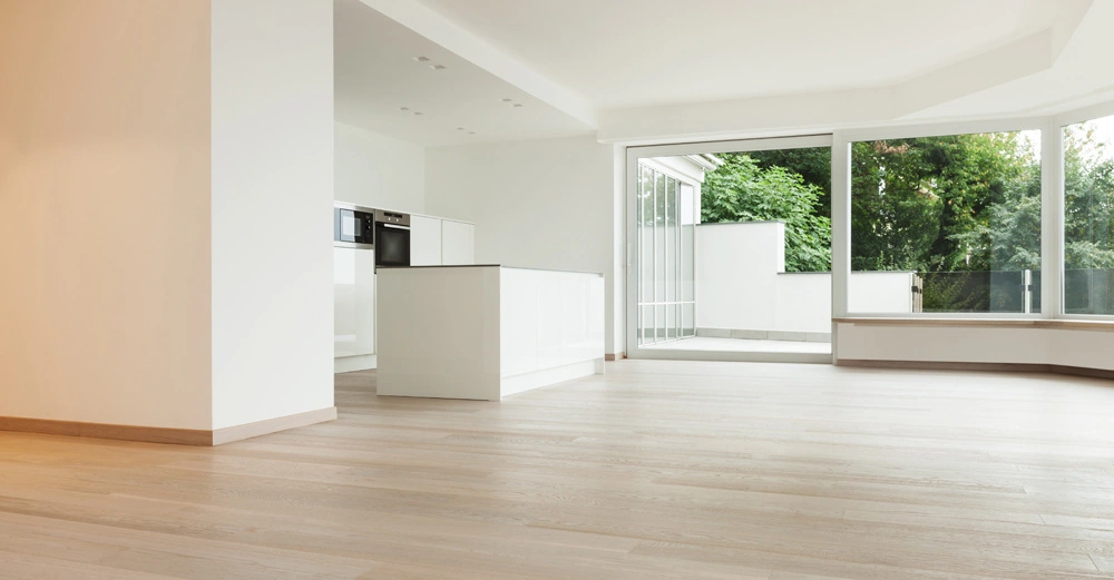 Geräumiges Wohnzimmer mit hellen Holzböden angrenzender moderner weißer Küche und großen Fenstern die Blick auf eine Terrasse und üppiges Grün bieten