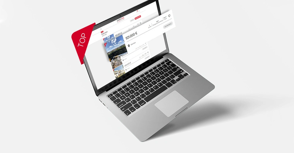 Ein Laptop zeigt eine Immobilien-Website mit Anzeigen für Häuser Preise beginnen bei 921.000 €. Ein roter Banner mit der Aufschrift TOP steht links. Der Laptop befindet sich auf einer weißen Oberfläche.