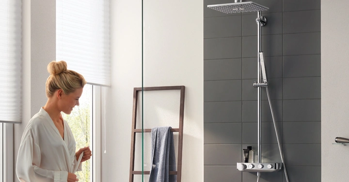 Eine Frau im Bademantel steht lächelnd vor einer modernen Dusche mit Kopfbrause in einem hellen, eleganten Badezimmer mit großen Fenstern und einem Handtuch auf einer Holzleiter.