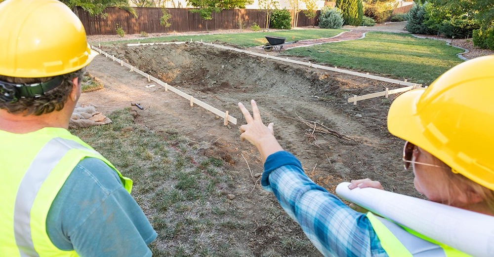 Arbeiter und Arbeiterin mit gelben Baustellenhelmen und Schutzwesten von hinten zeigen auf ausgehobene Baugrupe für Pool. Baustellensicherung mit Holzlatten vorhanden. Rest Gartenfläche