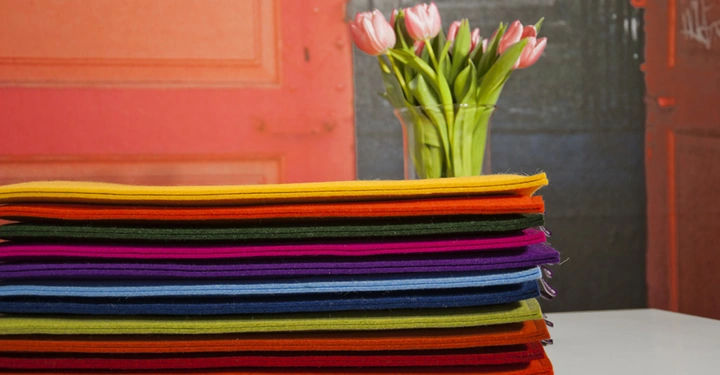 Galerie-Bild 7: Ein Stapel bunter Stoffbahnen liegt auf einem Tisch tulpenartige Blumen in einer Vase dahinter vor einem Hintergrund mit einer großen roten Tür.
